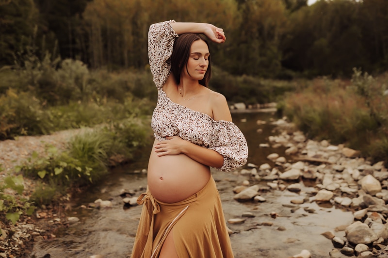 Maternity Photographer, an expectant mother places her hand on her belly before a quiet stream in the forest
