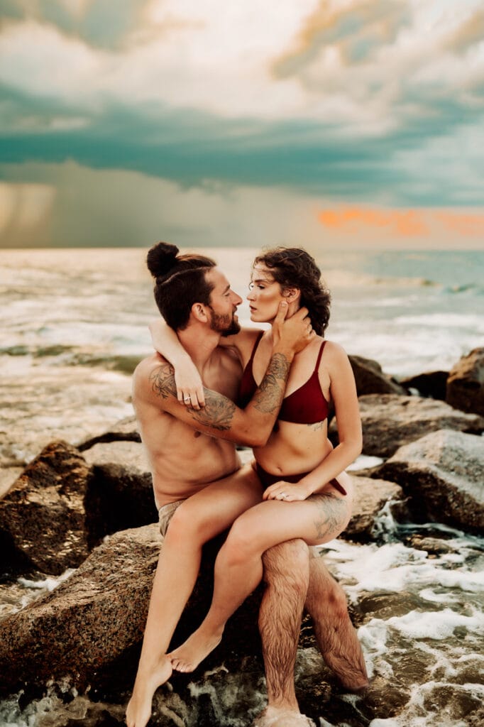 Family Photographer, a young couple sit on a rock near the ocean beneath the clouds, they are in the swimsuits, they gaze into each other's eyes