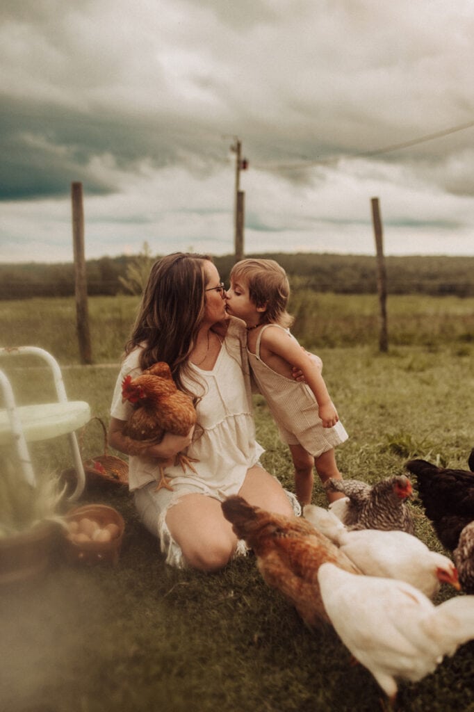 Family Photographer, a mother kisses her toddler as the chickens gather around them in the grass