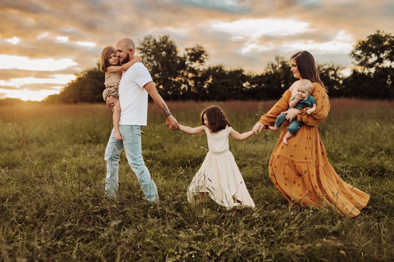 Newborn Photographer, a father carries a young daughter and leads his older daughter and wife through a field, she is a mother and holds their newborn baby