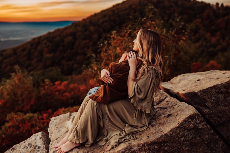 Family Photographer, a mother sits on a boulder overlooking mountain forests in the fall at sunset